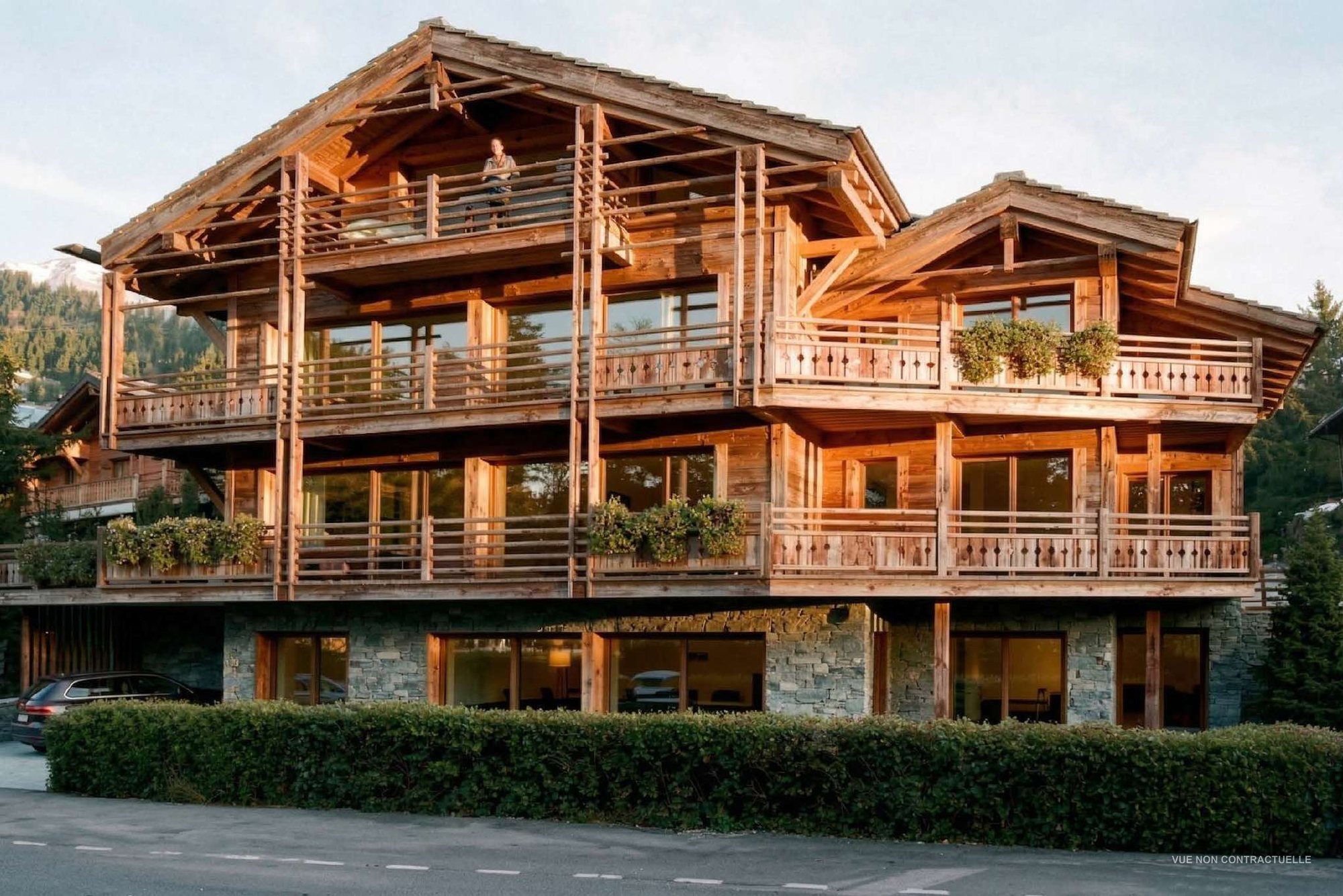 Vue de synthèse de la résidence Les Gentianes : un chalet contemporain en vieux bois sur socle de pierre, balcons fleuris, en fin de journée.